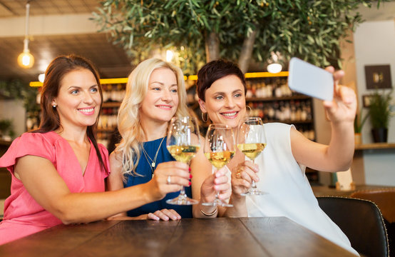 people, technology and lifestyle concept - women drinking wine and taking selfie by smartphone at bar or restaurant
