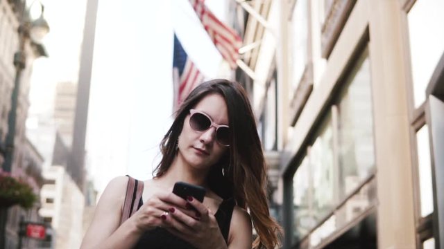 Happy Caucasian businesswoman in sunglasses using smartphone messenger app in New York street with USA flags slow motion
