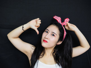Portrait of good looking sexy Chinese woman with long black hair, pink hair ribbon and white halter against black background, looking with thumb down gesture.