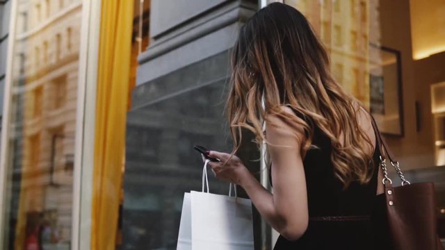 Camera follows young female beauty blogger carrying shopping bags in both hands walking along the street slow motion.
