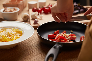 cropped shot of woman putting tomatoes and garlic into frying pan while cooking pasta