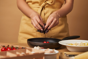 cropped shot of woman putting basil into frying pan while cooking pasta