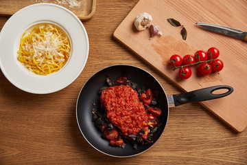 top view of plate of spaghetti and tomato sauce in frying pan on wooden table