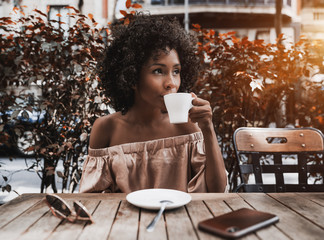 Beautiful African-American female is drinking delicious tea and looking aside while sitting in the...