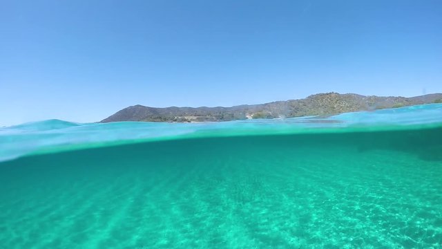 SLOW MOTION, HALF UNDERWATER: Picturesque view from the turquoise ocean of the distant island in the sunny Mediterranean. Cinematic half half of crystal clear sea and stunning Sardinia on a sunny day.