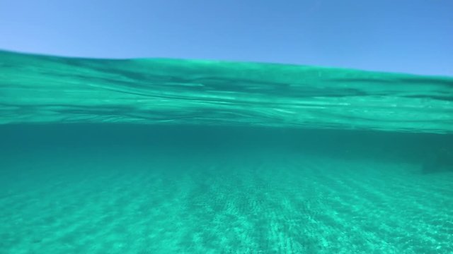 SLOW MOTION, HALF UNDERWATER: Spectacular shot of the distant island in the middle of the crystal clear ocean on a sunny day in summer. Awesome half underwater view of sandy ocean floor and Sardinia.