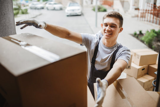 A young handsome smiling mover wearing uniform is reaching for the box while unloading the van full of boxes. House move, mover service.