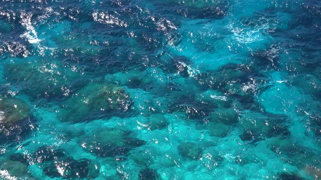 SLOW MOTION, AERIAL: Turquoise ocean water glimmering in the bright summer sun and splashing over the black rocks near the remote island coast. Flying above the rocky seaside in stunning sunny Italy.