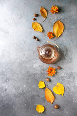 Glass teapot of hot tea decorated by yellow autumn leaves, aster flowers and acorns over grey texture background. Flat lay, space. Seasonal background.
