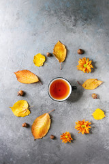 Cup of hot tea decorated by yellow autumn leaves, aster flowers and acorns over grey texture background. Flat lay, space. Seasonal background.