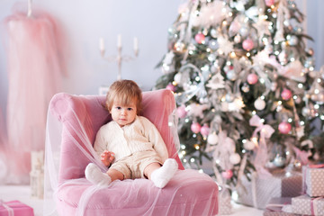 Baby boy 1 year old sitting in chair over Christmas tree at background. Looking at camera. Winter season.
