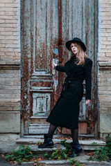 Blonde beautiful girl with long hair, in black coat in hat, stands on the background of vintage antique old wooden door old brick house. A beautiful girl tries to open a door. For concept design.
