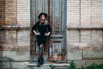 Fototapeta na wymiar Blonde beautiful girl with long hair, in black dress in hat, stands on the background of vintage antique old wooden door old brick house. For concept design.