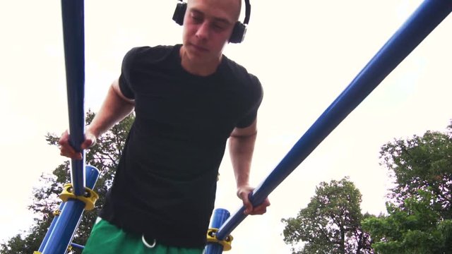 Slow Motion - Young Fit Man With Headphones is Engaged in Street Workout. Male Athletic Man Doing Triceps Dips on Parallel Bars. Summer Training in Park