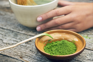 Matcha green tea powder in little clay bowl with girl's hand holding a big bowl with a finished tea on the background