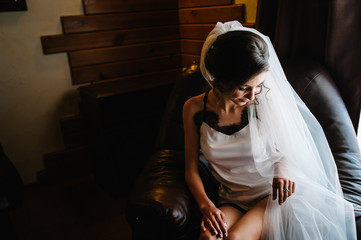 Naklejka premium The bride in sexy underwear or night dress from the veil on her head is sitting in the room on the armchair at home. Preparing for a wedding in the morning.