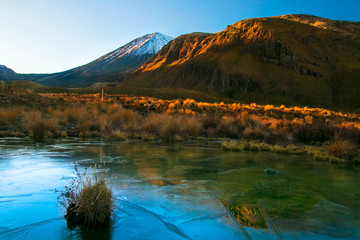 Early morning sunrise, landscape scenery of frozen blue pure water lake, wild mountains and huge volcano with the peak covered by first snow, autumn colours and golden sun rays, New Zealand nature