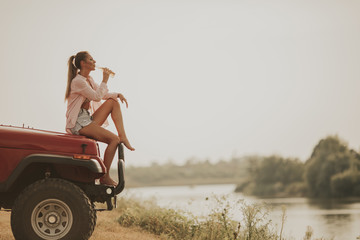Young woman sits on a car hood and drinks refreshment - obrazy, fototapety, plakaty