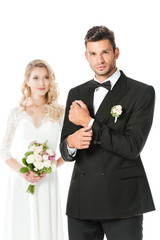 handsome young groom buttoning cufflink and looking at camera with bride standing blurred on background isolated on white