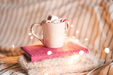 Cup of coffee with marshmallow and Christmas candy staying on book and knitted sweater over lights. Winter season.
