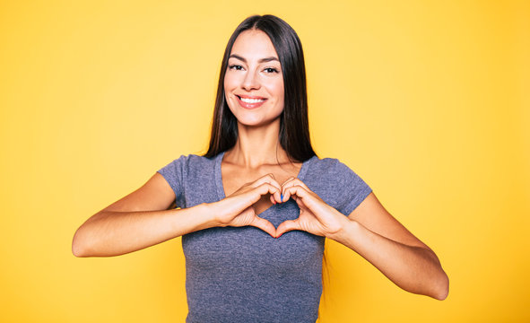 Love, heart shape, peace. Sign, gesture or symbols. Portrait of cute attractive smiling brunette woman, girl shows heart sign with help of her hands on yellow background in casual wear