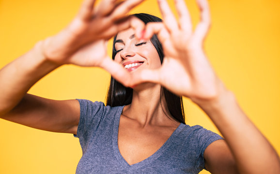 Love, heart shape, peace. Sign, gesture or symbols. Portrait of cute attractive smiling brunette woman, girl shows heart sign with help of her hands on yellow background in casual wear