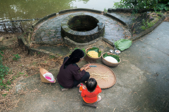 Grandmother and her granddaughter wrapping Chung Cake, the square shape cake, by old well and pond. Traditional Vietnamese New Year (Tet) food.
