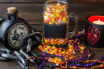 Halloween background frame with a jar of candy corn on a wooden table surrounded by a dark chain and next to a vial of poison and day of the dead candle with a black lable that you can personalize.