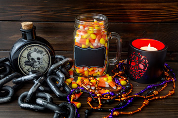 Halloween background frame with a jar of candy corn on a wooden table surrounded by a dark chain and next to a vial of poison and day of the dead candle with a black lable that you can personalize.