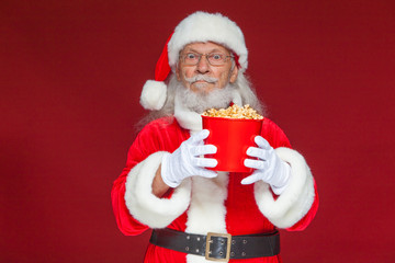 Christmas. Santa Claus in white gloves holding a red bucket of popcorn with two hands, moves it forward and offers to treat. The concept of visiting a cinema, watching a movie with popcorn. Isolated