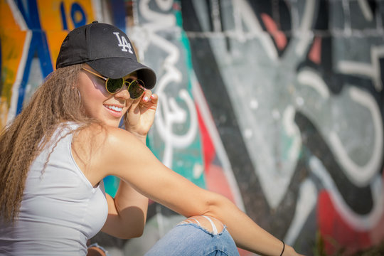 Portrait of young beautiful woman wearing white tank shirt and blue jeans and black hat on brick wall with graffiti background