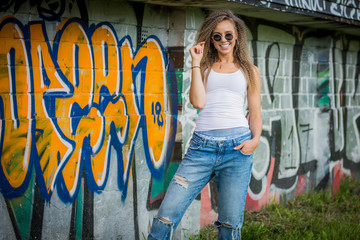 Portrait of young woman wearing white shirt and blue jeans and sunglasses on a brick wall with graffiti painting background