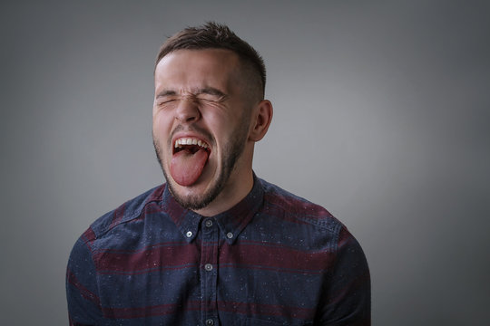 The adult man in a checkered shirt with a bristle on a beard wide open mouth puts out the tongue and closed eyes over gray background
