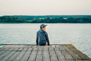 the little boy sits on the bridge near the lake. He is surrounded by a wonderful view