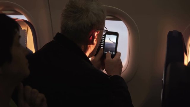 Unrecognizable senior male passenger taking smartphone photo of plane wing during flight sitting near airplane window.