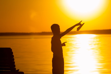 Fototapeta premium little boy stands on the background of the river at sunset. He holds a paper airplane in his hand and points his finger at the sun