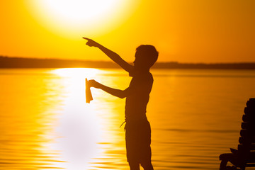 Fototapeta premium a little child stands on the background of the lake at sunset. In the left hand he holds a paper airplane, and with the right hand he points with his finger into the distance