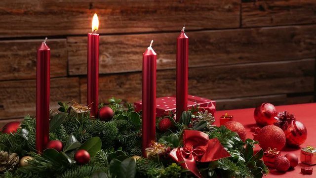 One burning red candle on a traditional advent wreath of green fir twigs and mistletoes with festive decoration in front of a rustic wooden wall, close-up real time shot with copy space, nobody, 30s