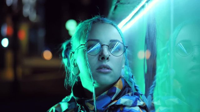 Young pretty girl with unusual hairstyle near glowing turquoise neon light of the city at night. Dyed blue hair in braids. Pensive sad hipster teenager in glasses