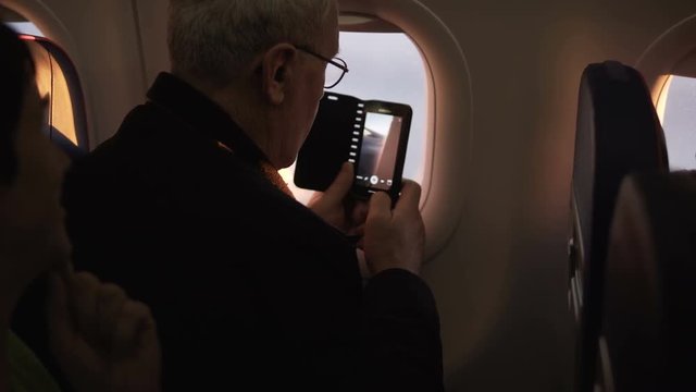 Unrecognizable senior male passenger trying to use smartphone camera during flight sitting near airplane window.
