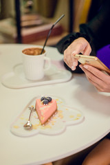 Women's hands with phone and pink cheesecake on a white dish-cloud, next to a mug of hot chocolate, the concept of a leisurely lunch in a cafe
