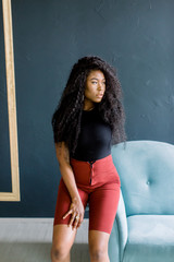Naklejka premium Young African American girl in black tshirt and red trousers healthy dark skin, poses indoor on sofa, has happy expression, expresses positive emotions. Facial expressions concept