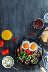 English Breakfast in a pan with fried eggs, sausages, bacon, mushrooms, jam and orange juice on dark stone background copy space, top view flatlay