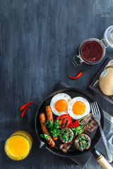 English Breakfast in a pan with fried eggs, sausages, bacon, mushrooms, jam and orange juice on dark stone background copy space, top view flatlay