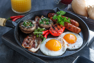 Close view of English Breakfast in a pan with fried eggs, sausages, bacon, mushrooms, jam and orange juice on dark stone background