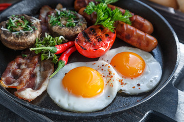 Close view of English Breakfast in a pan with fried eggs, sausages, bacon, mushrooms, jam and orange juice on dark stone background