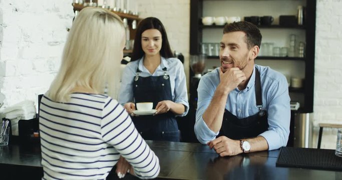 Attractive Caucasian waiter flirting with blonde woman at the bar, while pretty waitress serving her coffee.