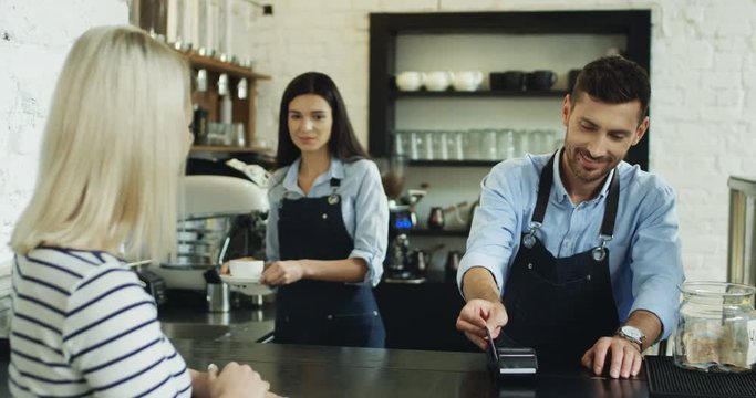 Back view on the blonde woman paying with a credit card to the waiter at the bar, while waitress serving her coffee.