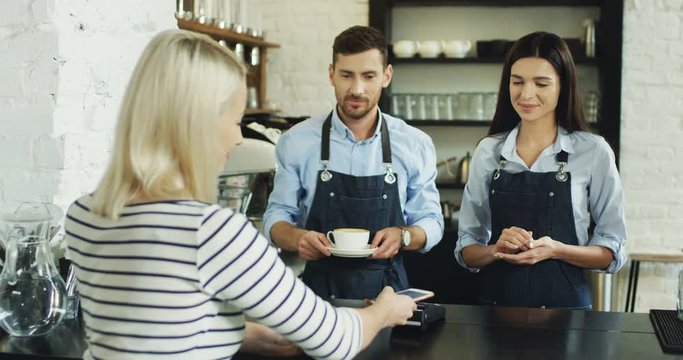 Rear of the blond woman paying with her smartphone at the bar while buying a coffee and waiter serving it. In the cafe.
