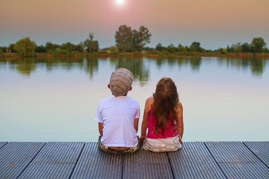 Boy with flat cap and little girl are sitting on pier. Boy and girl are looking upwards on sky. Love, friendship and childhood concept. Beautiful romantic sunset picture